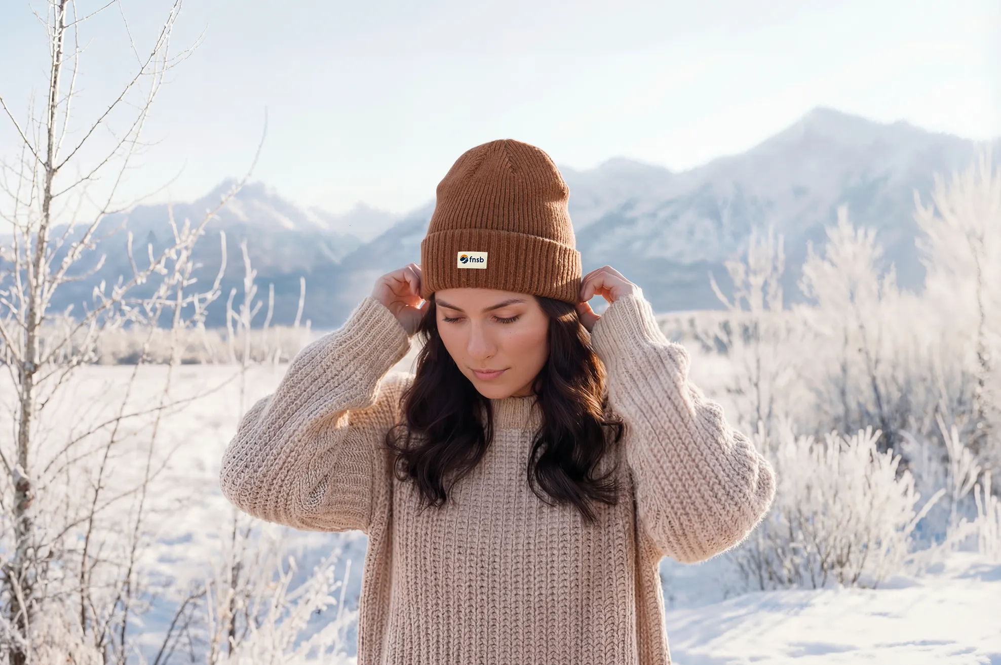 A person in a cream knit sweater pulls on a rust-brown toque with a small FNSB logo label, standing in a snowy Yukon field with frost-covered trees and mountains behind them.
