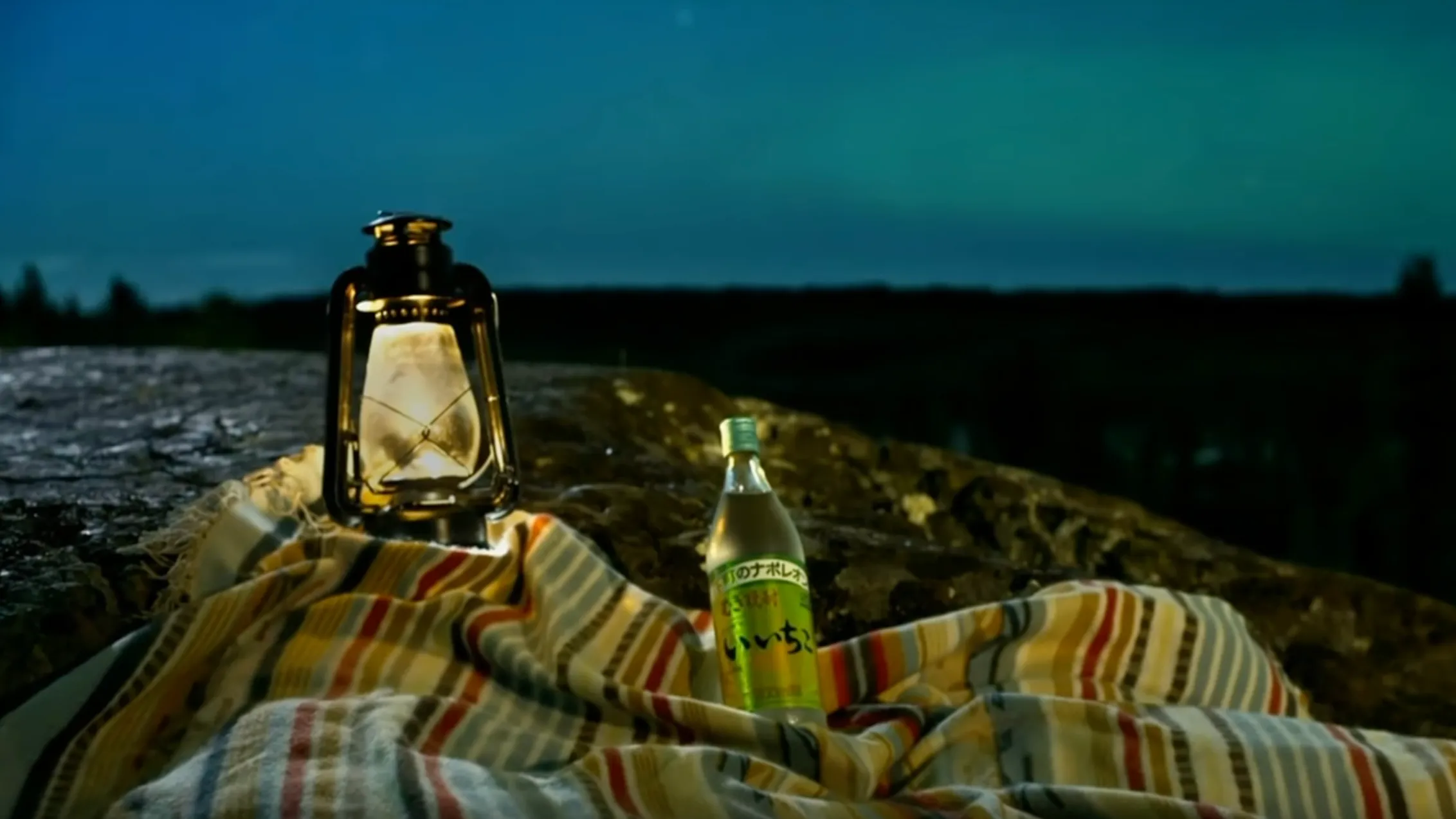 Cinematic lifestyle product photography of a bottle of iichiko shochu and a lit vintage lantern on a blanket outdoors at dusk.